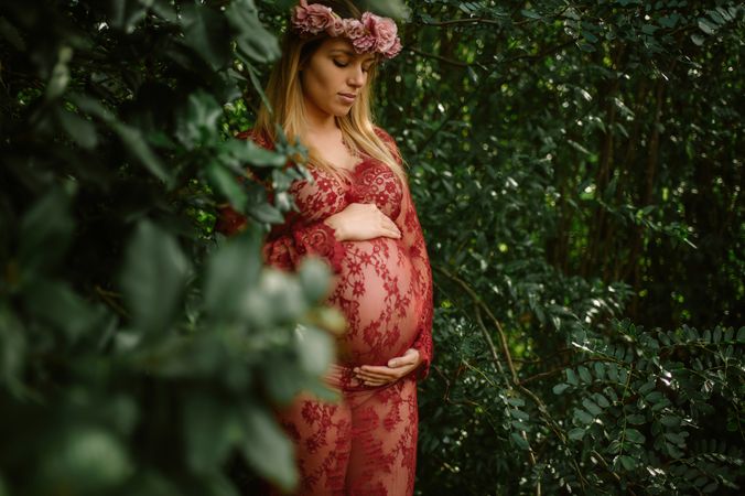 Pregnant woman with closed eyes touching belly and chin while standing in garden in sunny day