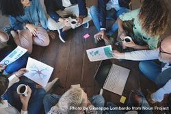 Aerial view of colleagues sitting on wooden floor during a brainstorming session, sharing coffee, laptops, and documents with charts and mind maps bGRNla