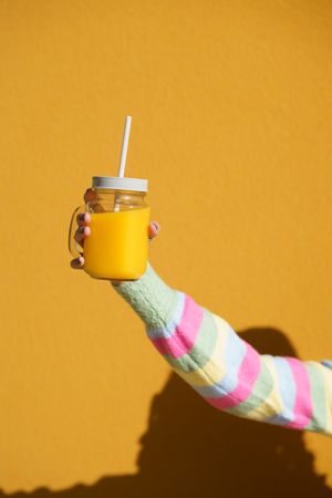 Vertical shot of person in striped sleeve holding cup of orange juice on yellow wall
