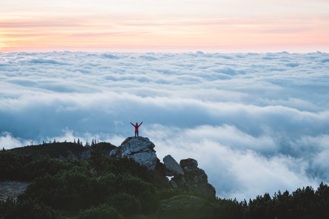 Person standing on rock raising hand on top of the mountain above the clouds
