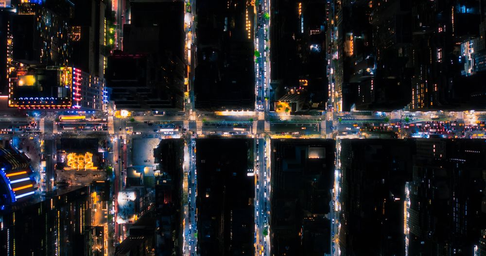 Top Down Aerial View of New York City Streets at Night with Visible Grid System, Business and Residential Building Roofs. Busy Center with Traffic, Cars, Yellow Taxis, Commercial Vehicles, Pedestrians