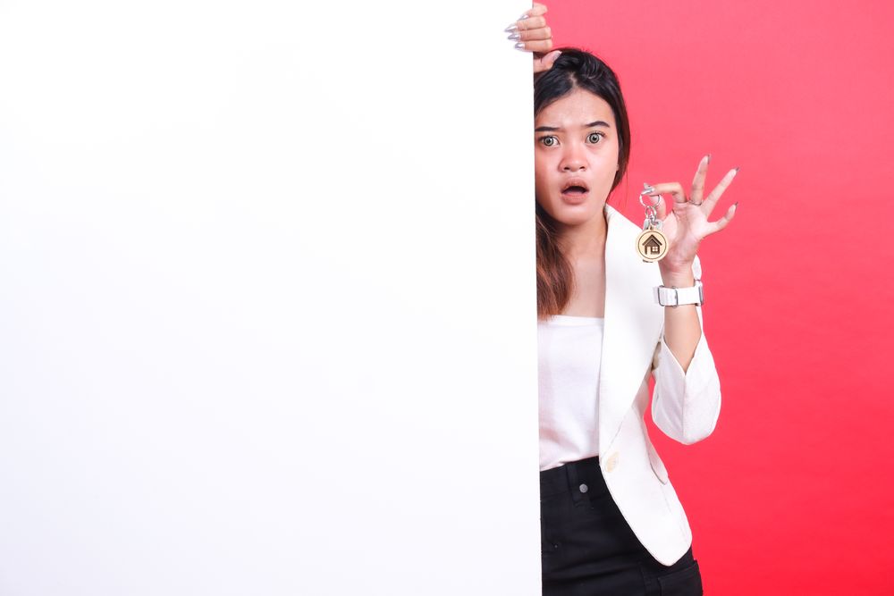 A woman is holding a house key and hiding behind a banner with a surprised expression, isolated on a red background