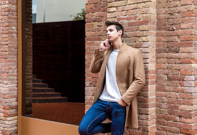 Male in camel coat talking on phone while leaning back on brick wall