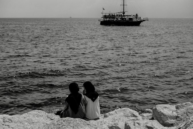 Back view of two women wearing the hijab sitting on rocky beach in grayscale
