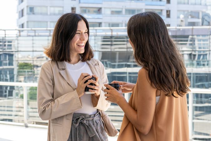 Two businesswomen are standing outside a building, enjoying their office break. They are smiling and holding cups of coffee, indicating a sense of camaraderie and friendship.