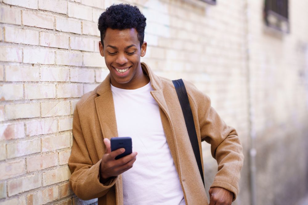 Happy Cuban male leaning against wall looking down at his phone