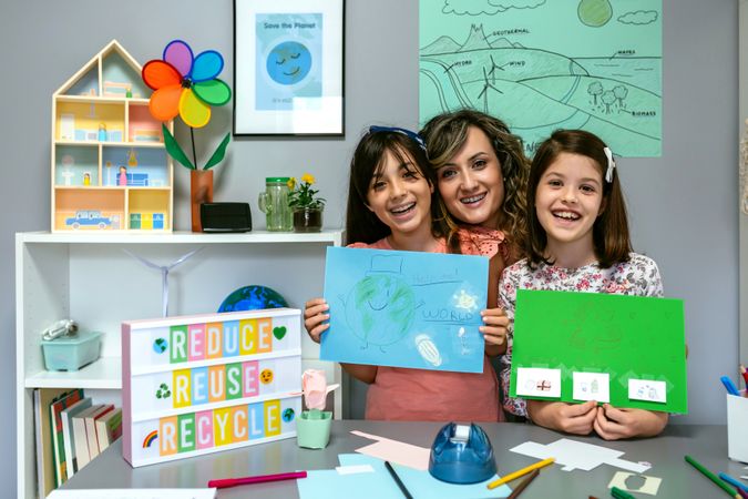 Teacher in classroom looking at camera next to students showing colorful ecological drawings