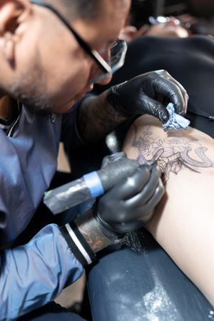 A tattoo artist is tattooing a snake and a rose on a woman's leg. Close up.