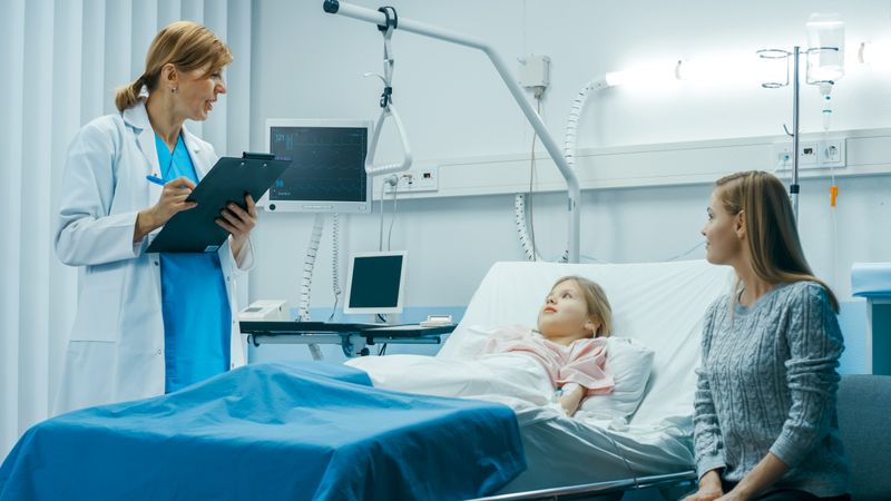 Sick Little Girl Lies on a Bed In the Hospital, Friendly Doctor With Clipboard Asks Where it Hurts, Mother Sits Beside Bed