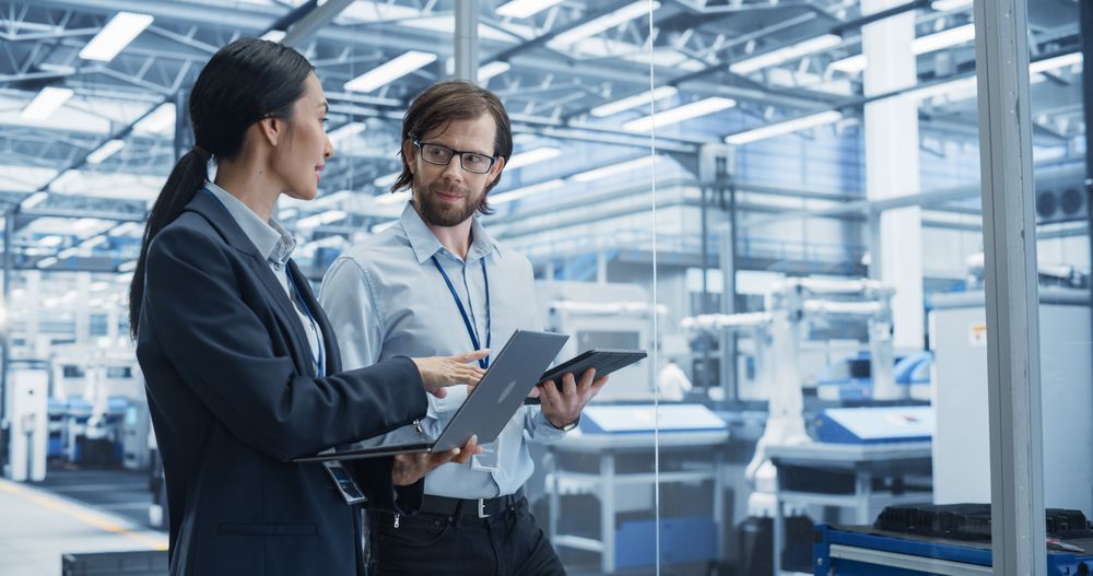 Portrait of a Cheerful Engineer and Happy Asian Scientist Using Laptop and Tablet Computers, Talking in a Factory Facility with Robots Producing Modern Electronic Components for Different Industries