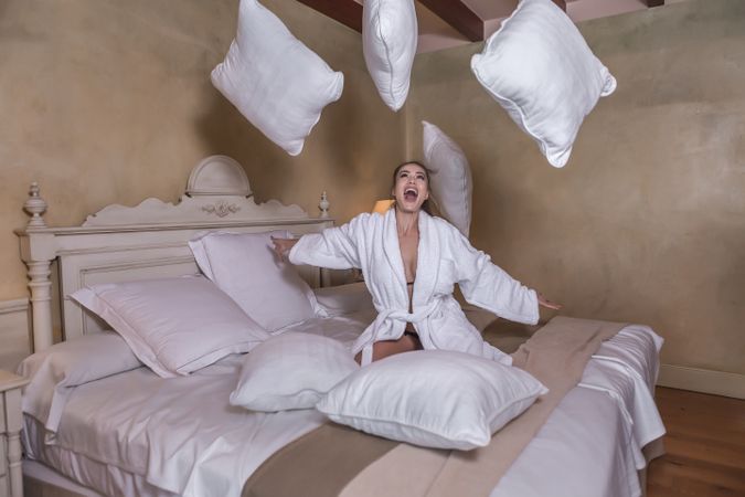 Excited woman in bathrobe throwing pillows in a hotel room