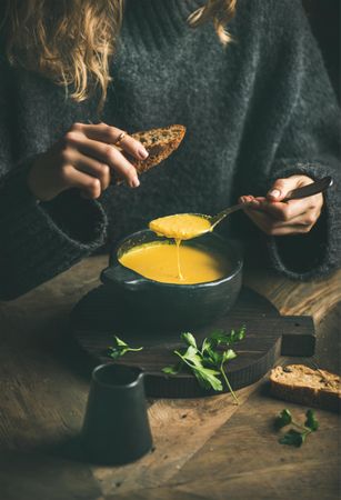 Blonde woman eating warm yellow soup from dark bowl holding toast