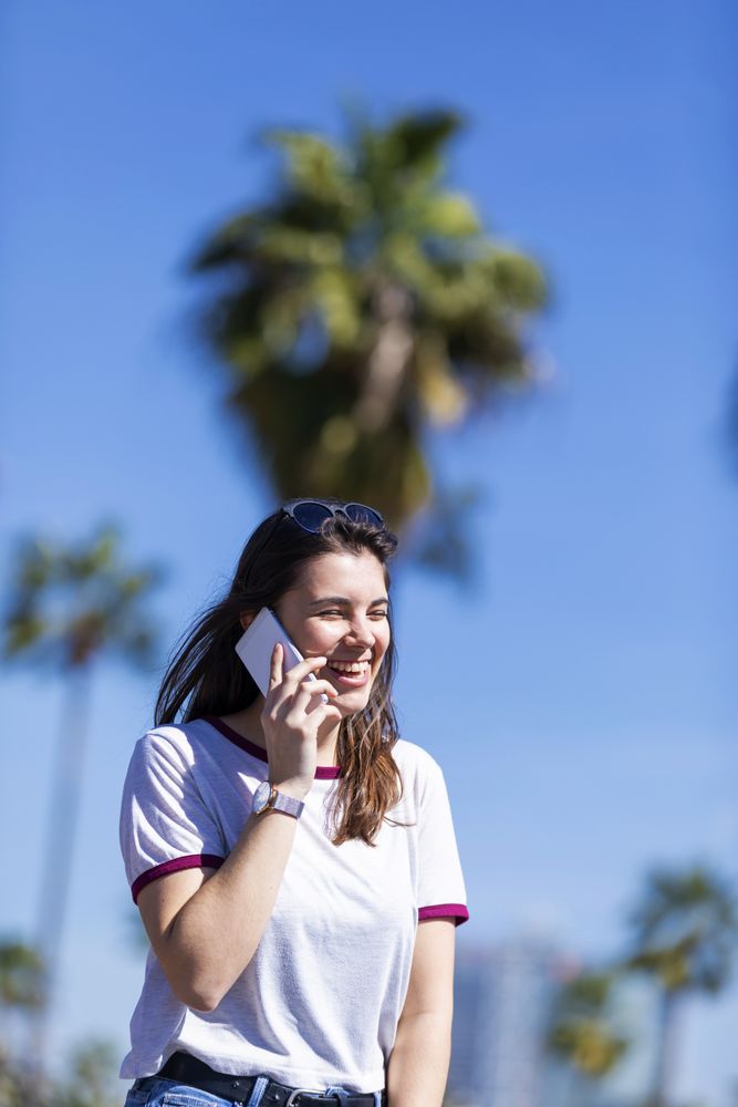Laughing woman wearing clothes standing outdoors in the street while using a mobile phone on a sunny day