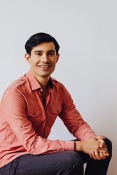Male sitting in grey studio smiling in pink shirt