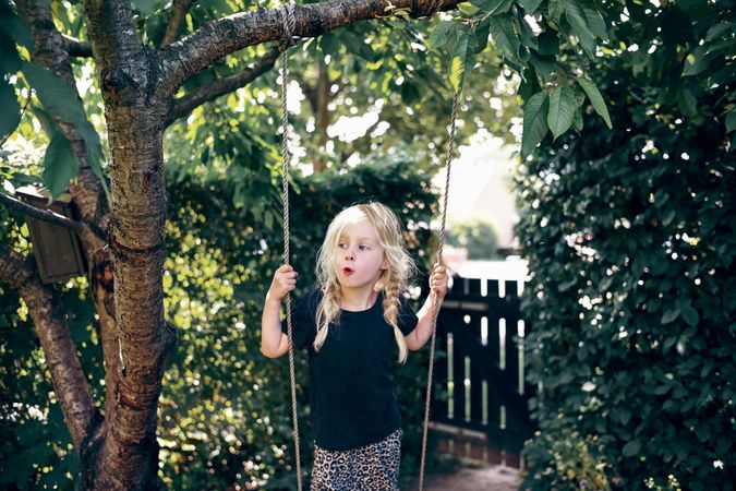 Surprised blonde girl with braided hair standing on outdoor swing in backyard