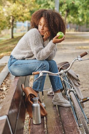 Pensive Black Latina woman sitting on bench in park with apple
