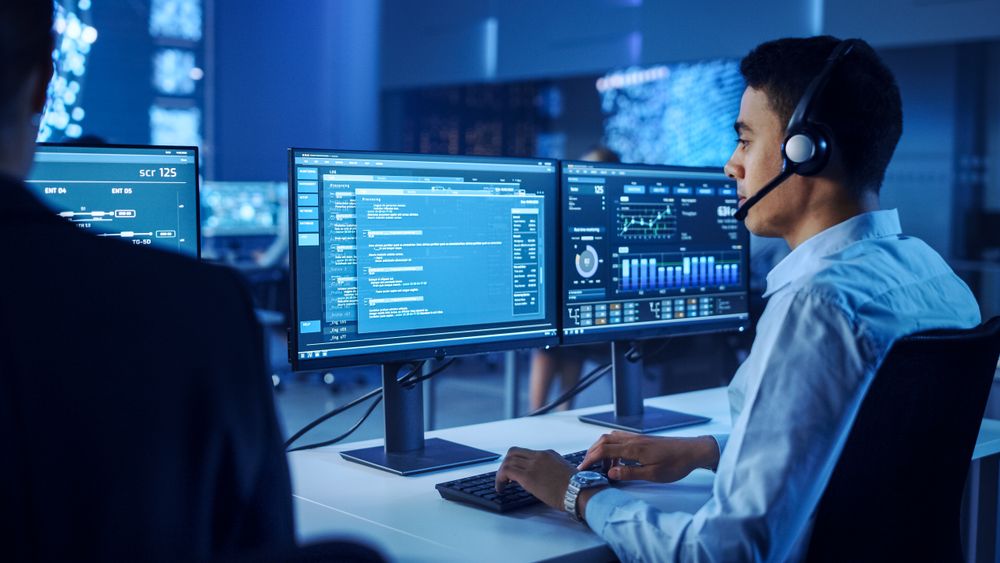 Professional Male Monitors Cybersecurity Data on Dual Screens in a High-tech Control Room, Optimizing Strategies With Precision and Innovation.