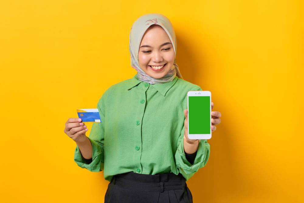 Happy Muslim woman in headscarf and green blouse holding credit card and smart phone screen mock up