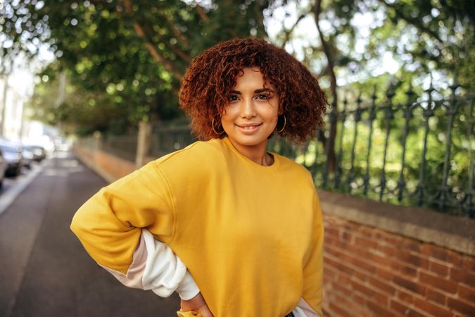 Confident biracial young woman standing on sidewalk in front of a park smiling and looking at camera