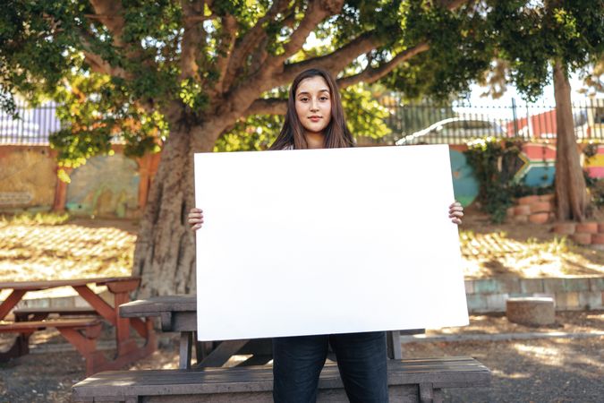 Confident teenage girl looking at the camera while holding a blank poster in an urban park