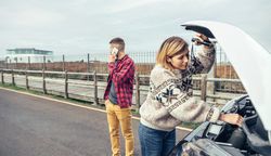 Female in woolen sweater working on car engine while partner makes emergency call