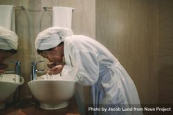 Woman in bathrobe washing her face in bathroom sink 0WPG65
