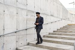 Young Latino gay man wearing a fashionable hat and sunglasses, walking down the stairway while texting on his smartphone