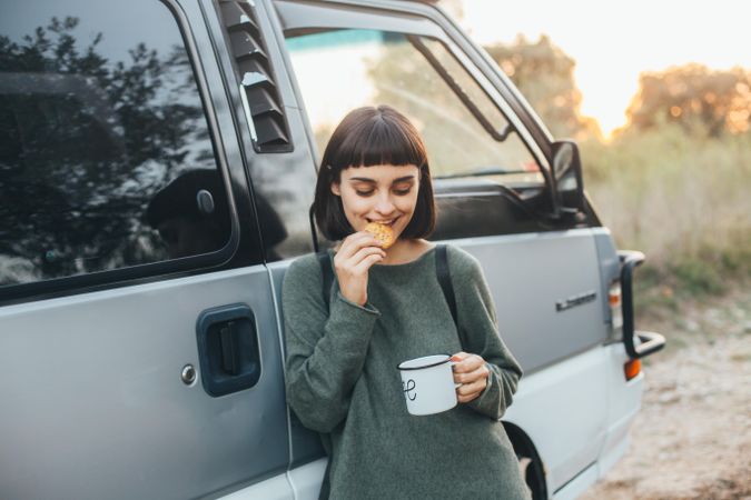 Woman with dark hair eating a cookie with coffee next to a van