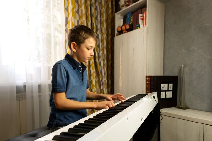 Side view of boy playing digital piano near bookshelf