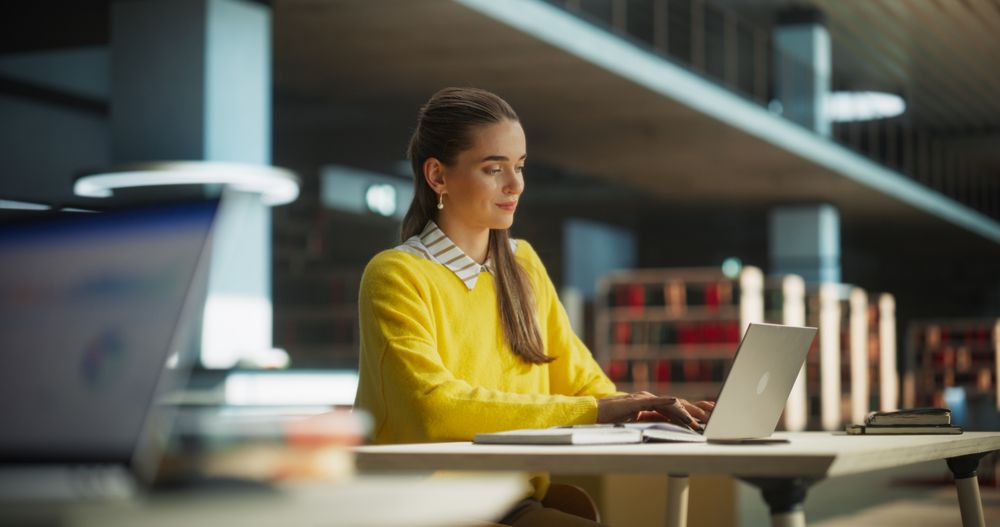 Female Student in Yellow Jumper Working on Her School Thesis on a Laptop Computer. Young Smart Woman Studying Online in a Quiet Public Library, Writing Down Report in Notebook