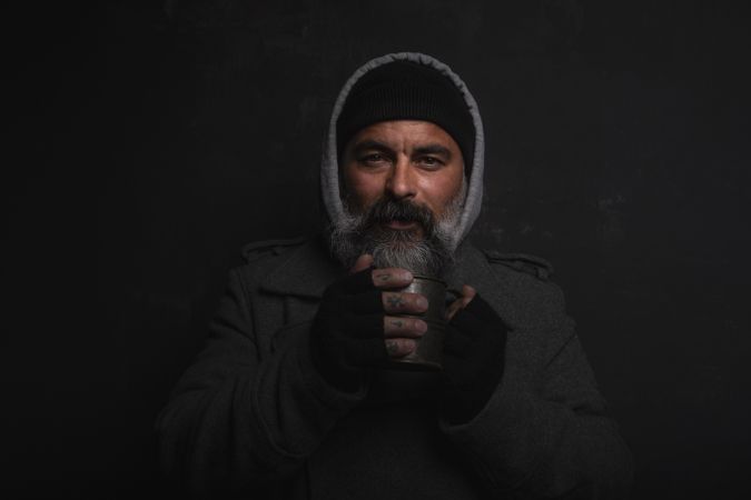Homeless middle-aged man with a gray beard holding a cup of hot tea against dark background