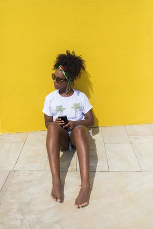 Portrait of Black woman in t-shirt sitting with phone while leaning on a yellow wall