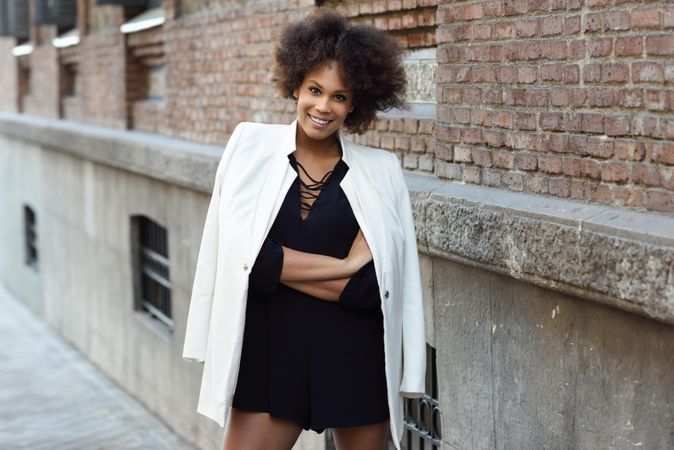 Smiling female in dress with blazer draped over shoulders standing next to outdoor wall, copy space