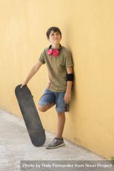 Young boy leaning on a yellow wall with headphones on neck, holding a skateboard while looking up bYqzkg