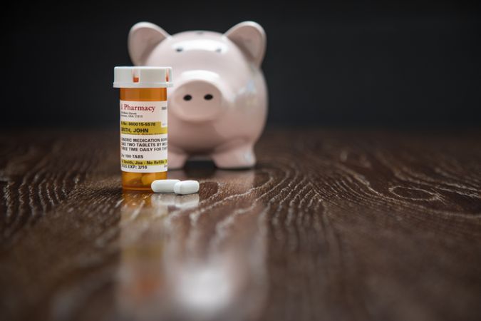 Non-Proprietary Prescription Medicine Bottle, Pills and Piggy Bank on Reflective Wooden Surface.