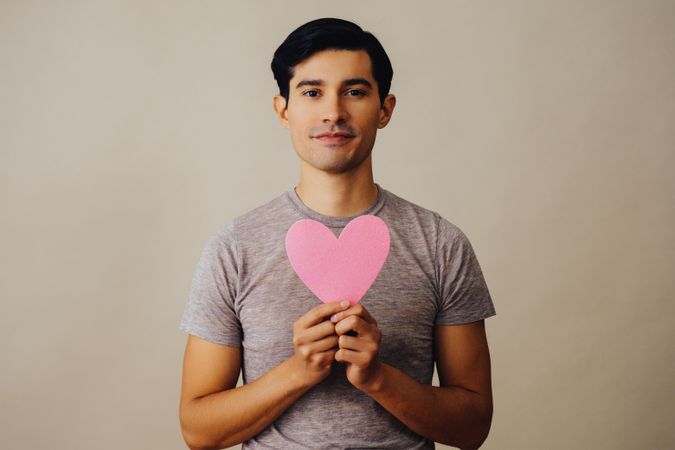 Romantic Hispanic male holding cut out pink heart to his chest