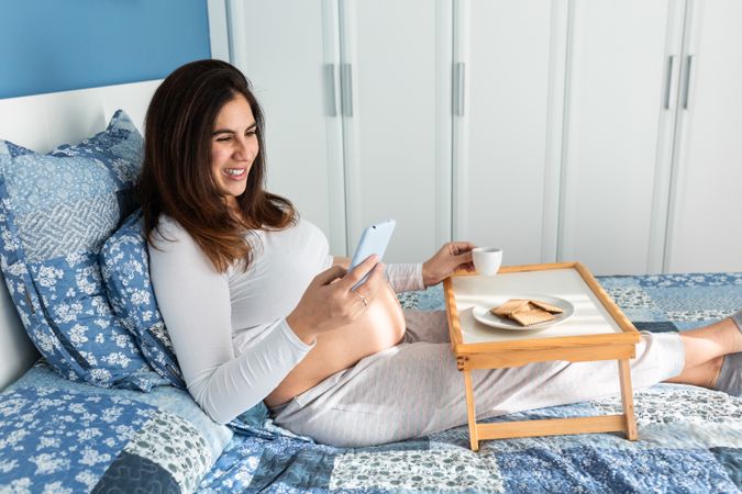 Happy pregnant woman having breakfast and browsing smartphone