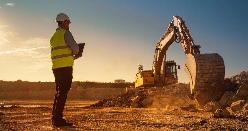 Male Civil Engineer Using Laptop Computer On Construction Site Of New Real Estate Project. Man Observing Industrial Heavy Machinery At Work, Laying Foundation For Apartment Building. Sunny