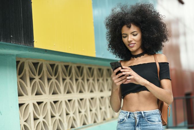 Young female with afro hair checking phone near a modern colorful building