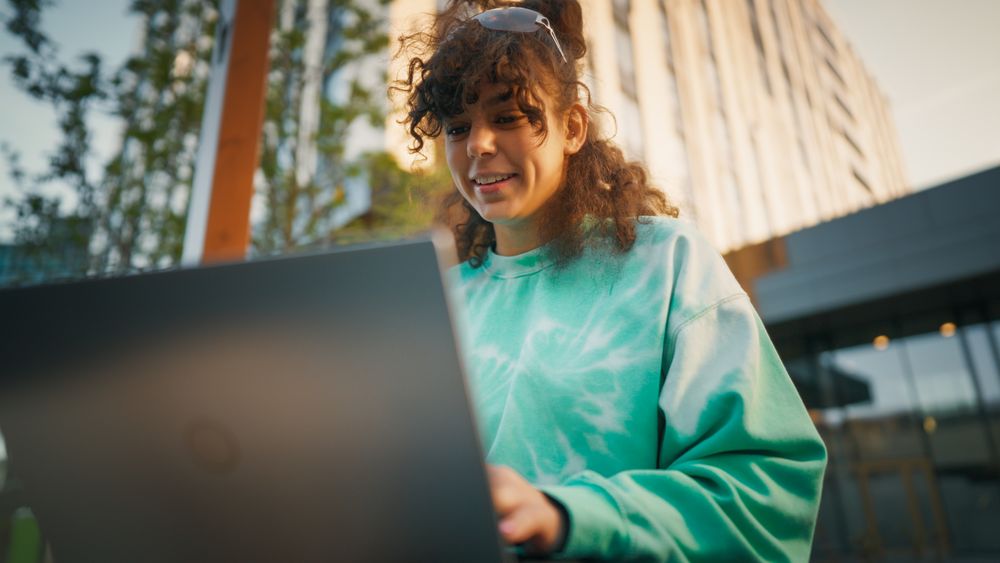 Young Woman With Curly Hair, Wearing A Turquoise Sweatshirt, Engaging With A Laptop Outdoors