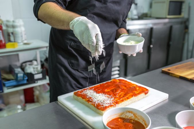 Unrecognizable male chef sprinkling grated cheese over tomato sauced focaccia in restaurant kitchen