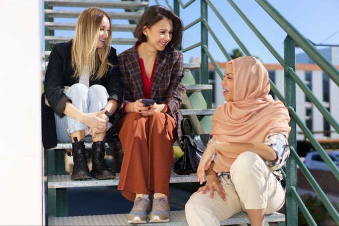 Trendy multi-ethnic women friends in stylish outfits sitting on metal stairs