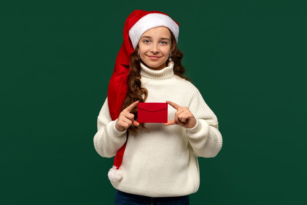 Girl in Santa hat holding red holiday envelope with smile.
