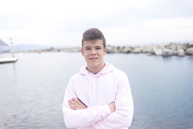Young male teenager with crossing arms standing on the coast while looking at camera