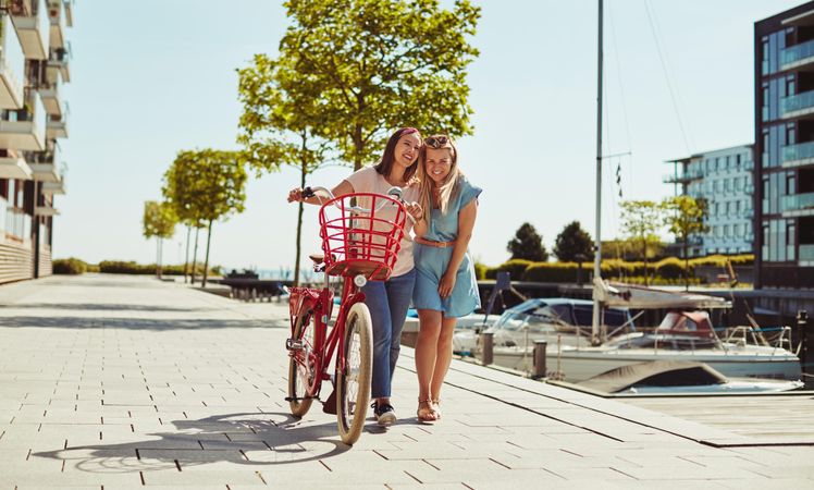 Two happy women walking with a bicycle along the river walk