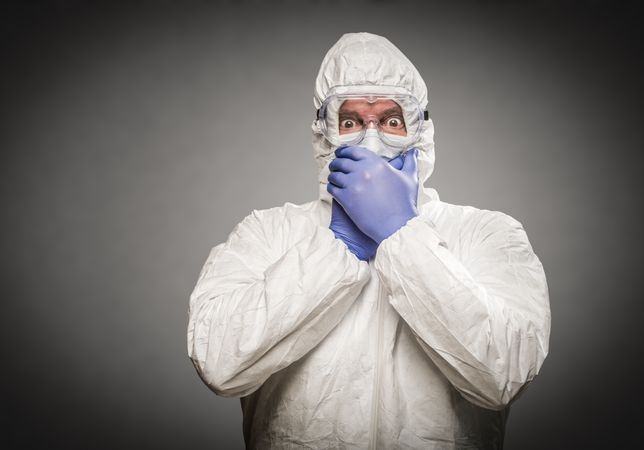 Man Covering Mouth With Hands Wearing HAZMAT Protective Clothing Against A Gray Background.