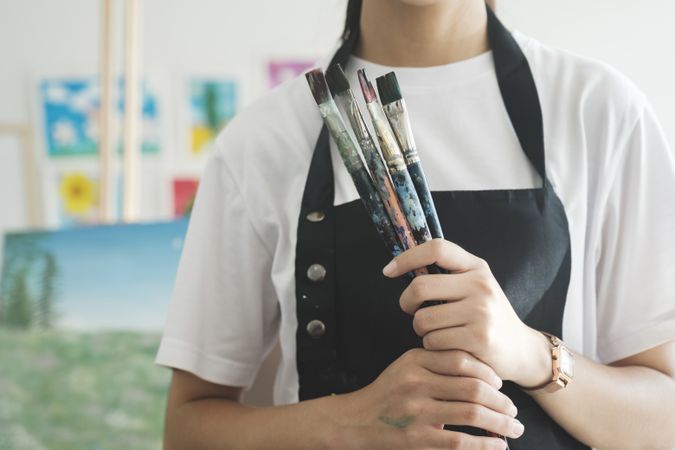 A close-up of the artist hands wearing an apron smeared with paint. Clutching many brushes and paintbrushes.
