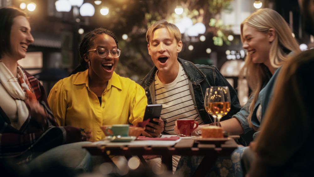 Young Female Sitting on a Terrace with Multiethnic Friends, Showing Holiday Photos on a Smartphone.