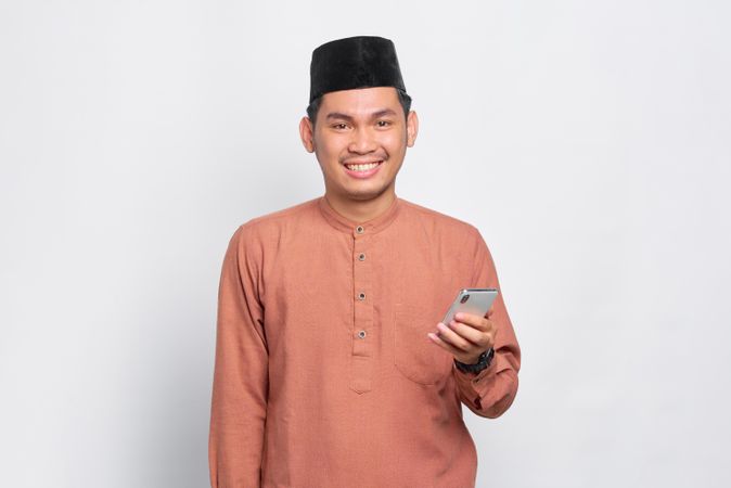 Happy Muslim man in kufi hat smiling and holding mobile phone