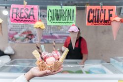 A hand is holding a traditional Mexican decorated ice cream in front of a street market stall with flavor signs